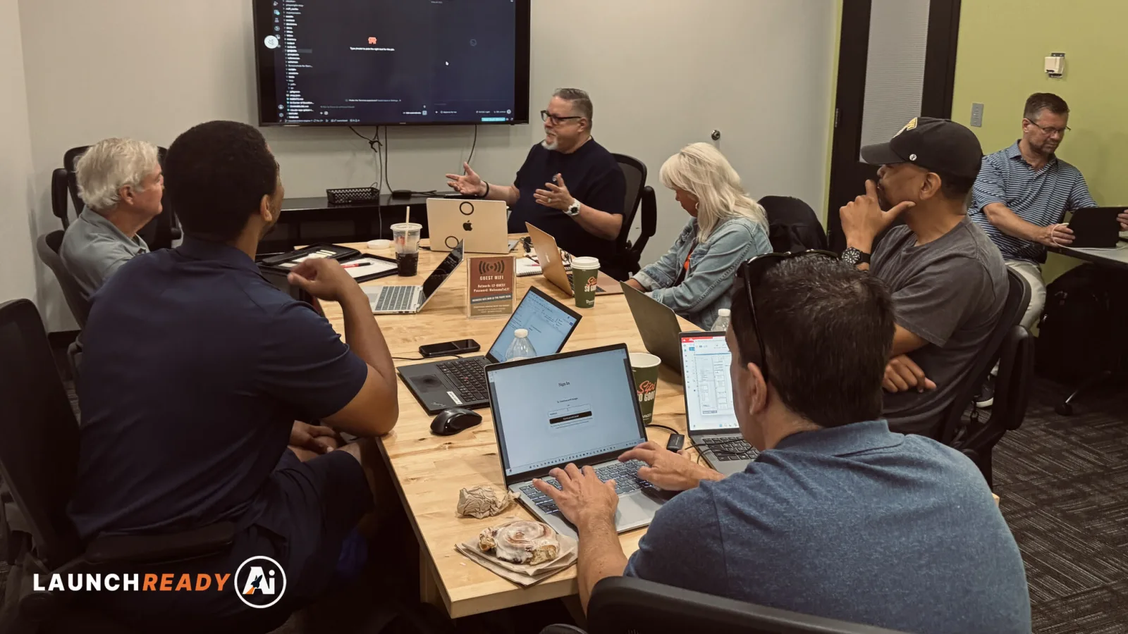 Eight people around one table at a LaunchReady AI workshop in the Indianapolis area, laptops open, Harrison Painter talking them through a step on the wall screen.