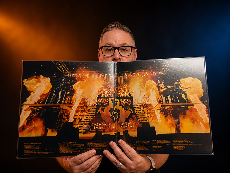 Harrison Painter holding the open KISS Alive II gatefold album, with the band-on-stage fire imagery dramatically lit by orange and blue stage-style lighting